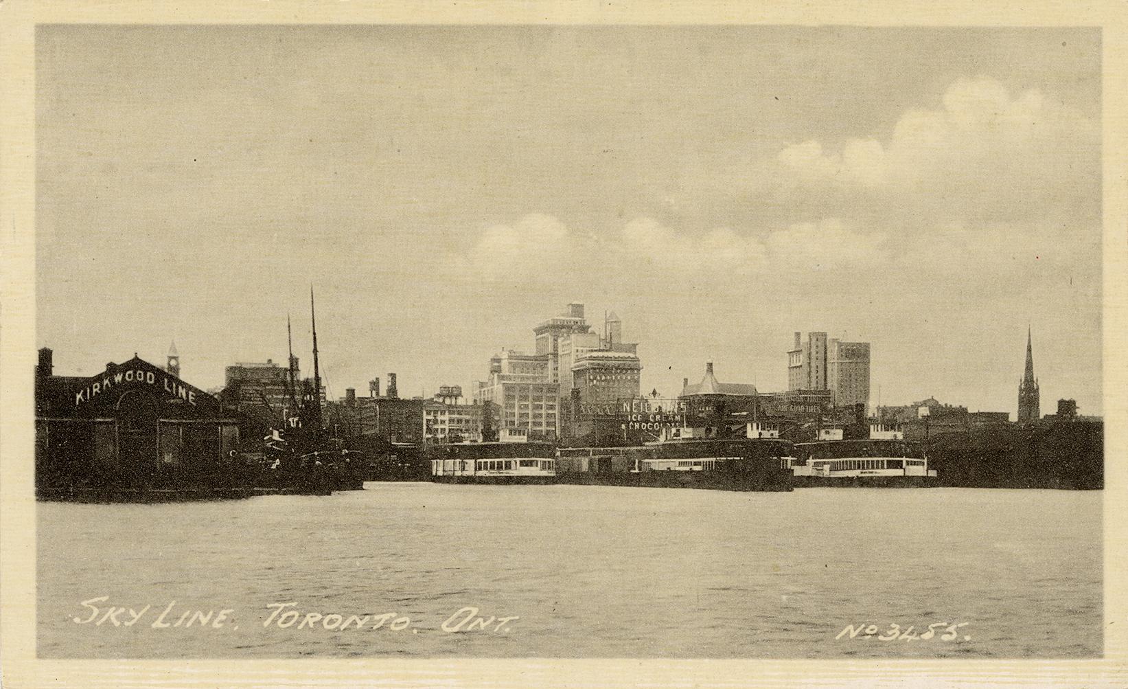 Black and white photograph of the shoreline of a large city. Docks and boats in the foreground.