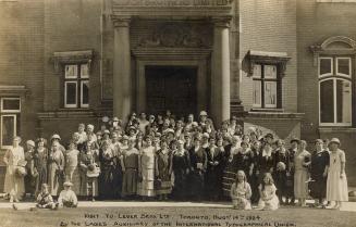 Black and white photograph of a large group of woman posing in front of the front entrance of a…