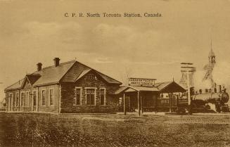 Sepia toned photograph of a public building with a train engine to the right of it. 