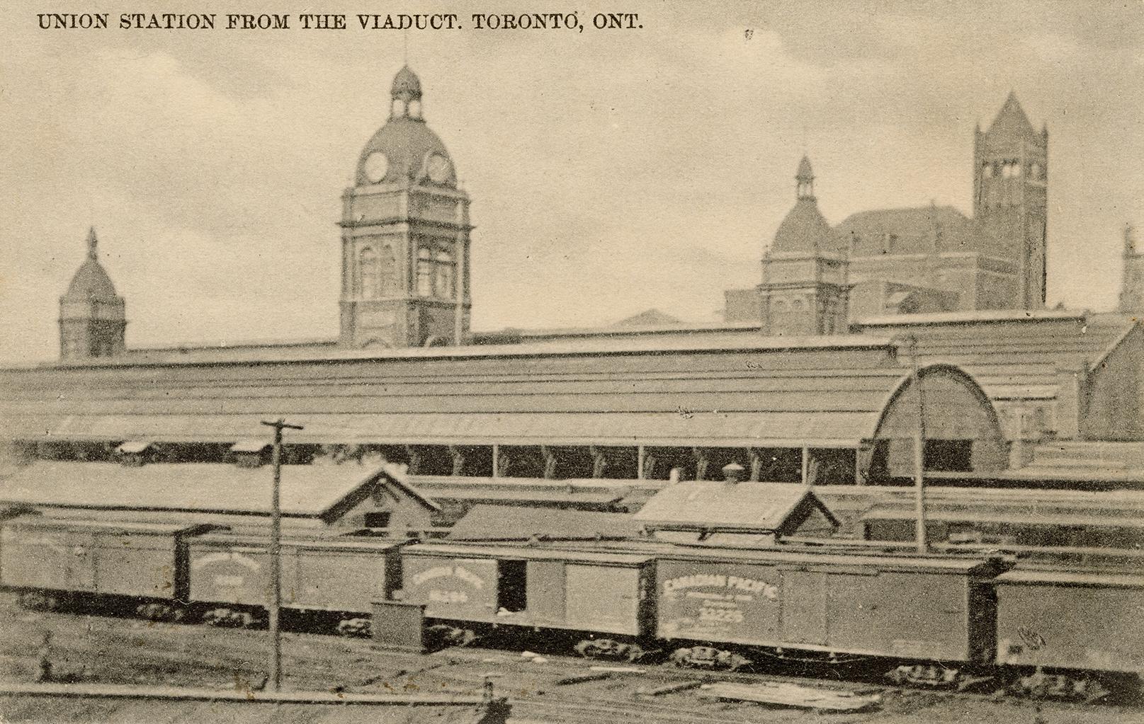 Black and white photograph of railway box cars on rails in front of terminal buildings.