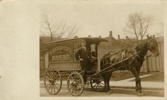 Black and white photograph of a man proudly standing on his horse drawn wagon.