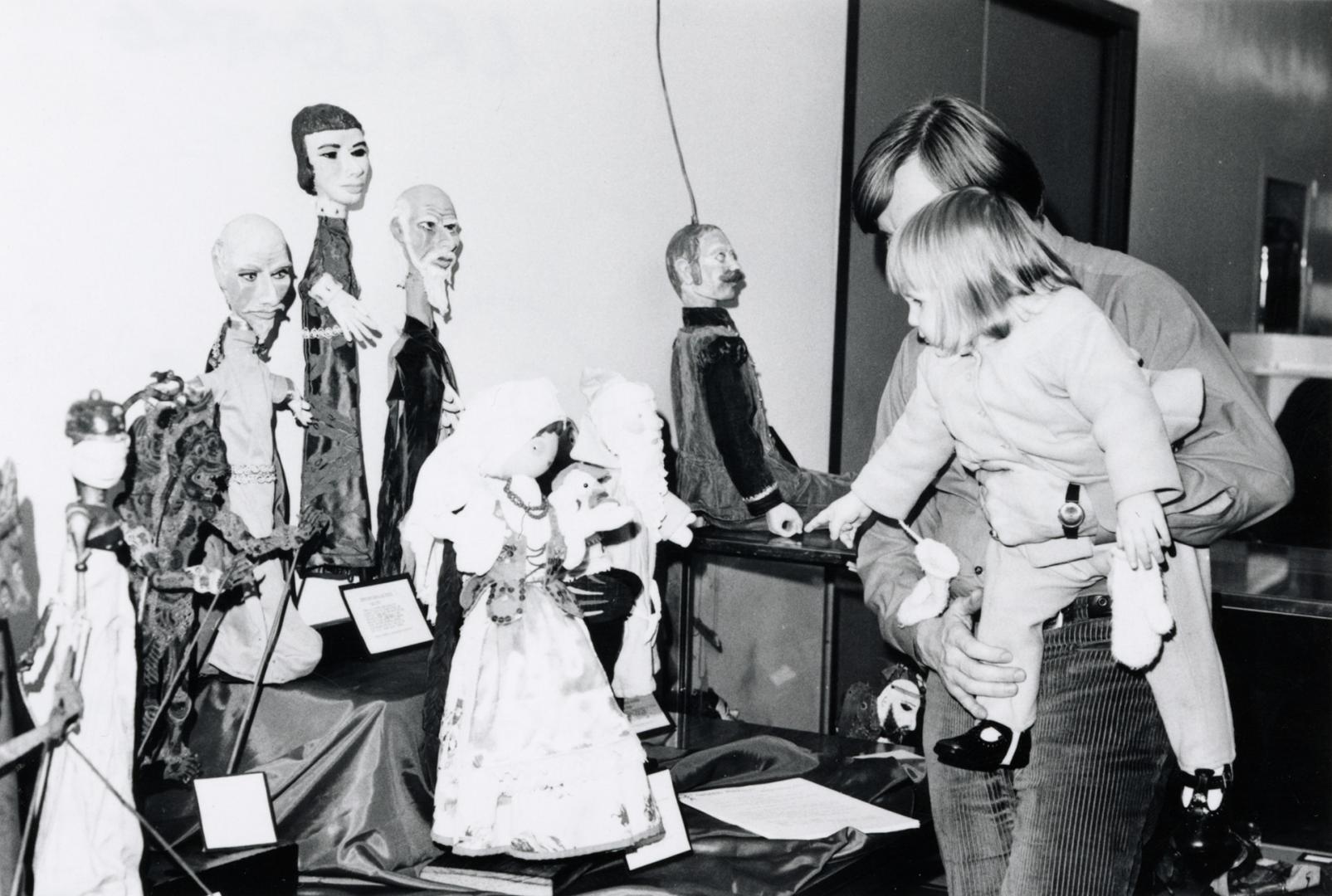 Picture of a man holding a child and looking at a display of puppets on a table. 
