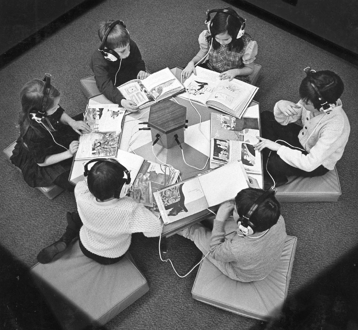 Six children sit around a table looking at books and wearing headphones to listen to an audio r ...