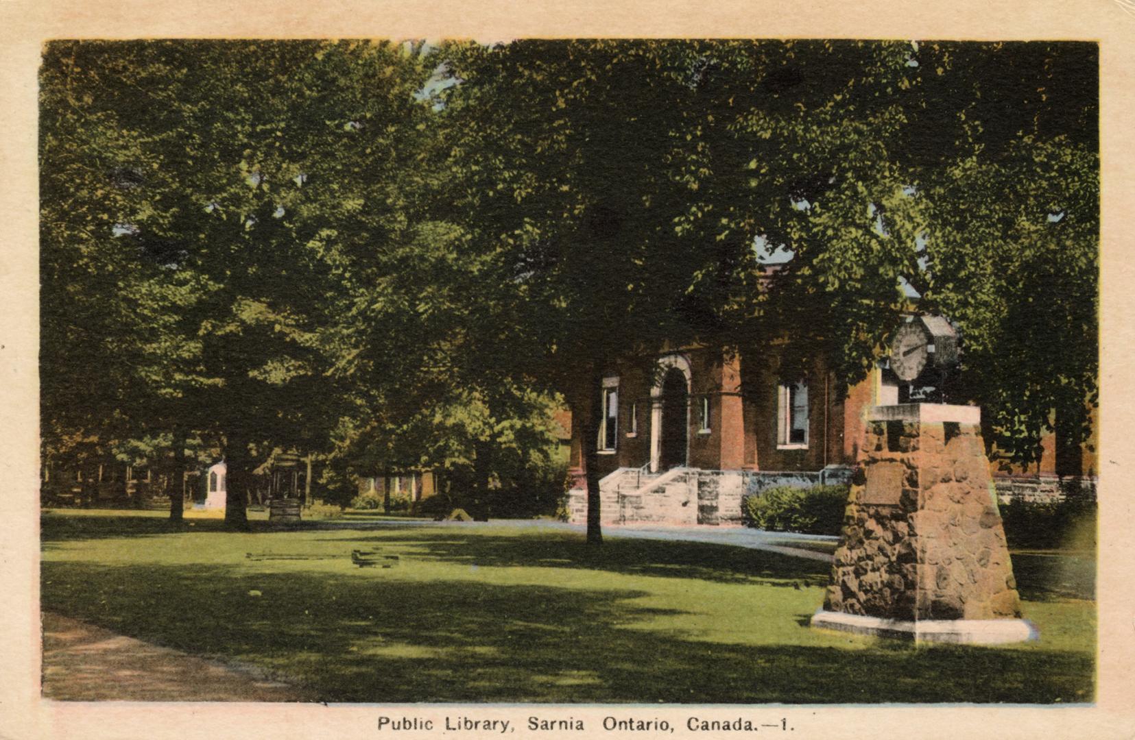 Picture of a brick library building mostly hidden by trees with park and stone memorial in fron ...