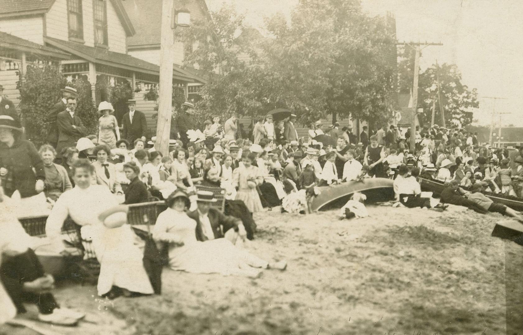 Black and white photograph of a huge crowd of people with canoes sitting on a shoreline.