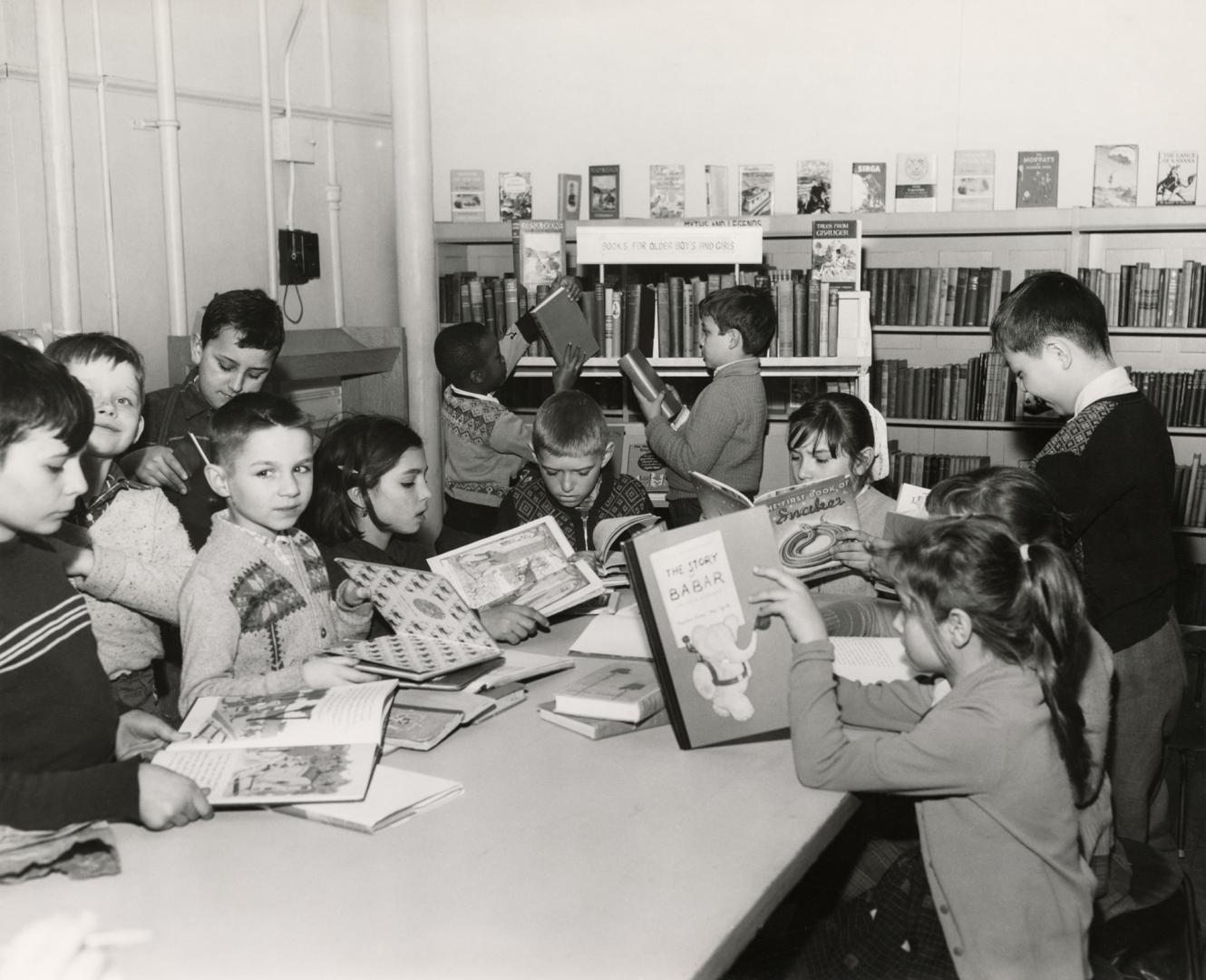 Picture of a group of boys and girls reading books at a large table while others look at books  ...