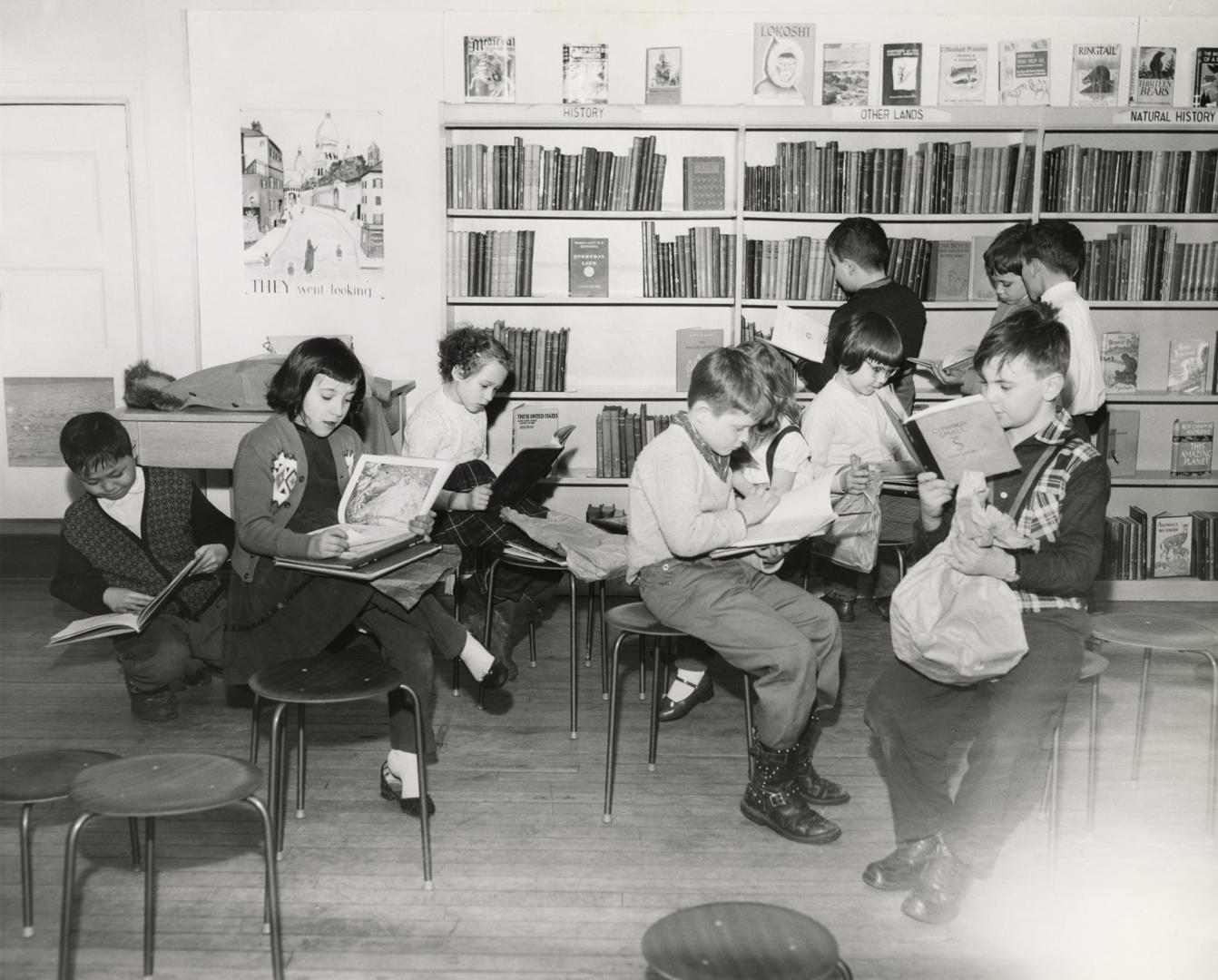 Picture of a group of boys and girls reading books while sitting on stools while others look at ...