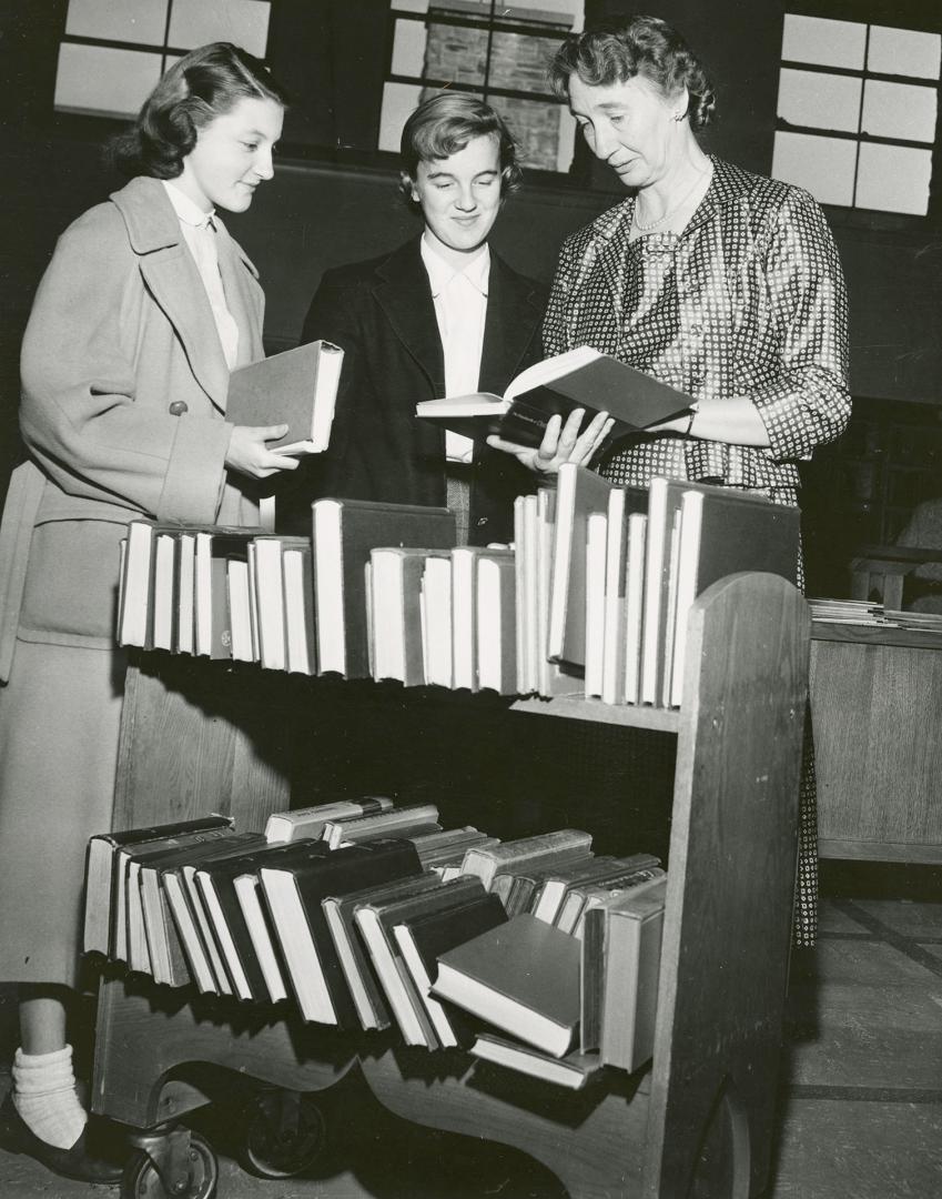 Picture of librarian standing at a book truck showing a book to two teenager girls. 