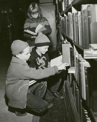 Picture of librarian standing at a book truck showing a book to two teenager girls. 