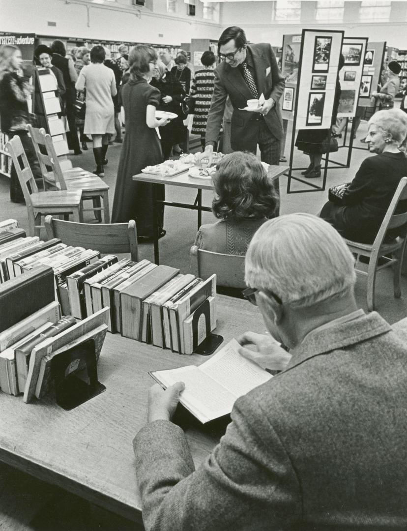 Picture of man reading at a desk in a library and people in background looking at a display. 