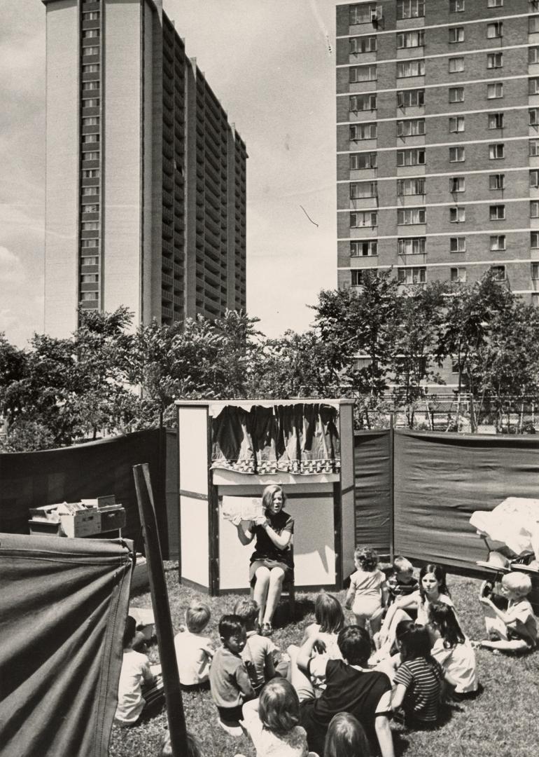 Picture of children sitting outside listening to a librarian storyteller in a park with apartme ...