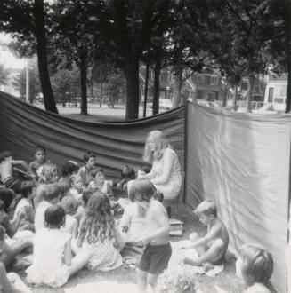 Picture of a group of children in a park listening to a story from a librarian. 