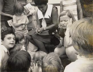 Picture of a group of children in a park listening to a story from a librarian. 