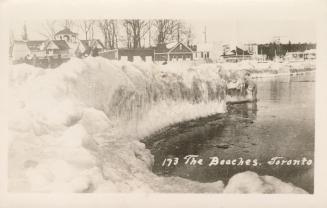 Black and white photograph of a frozen beach shoreline with houses in the background.