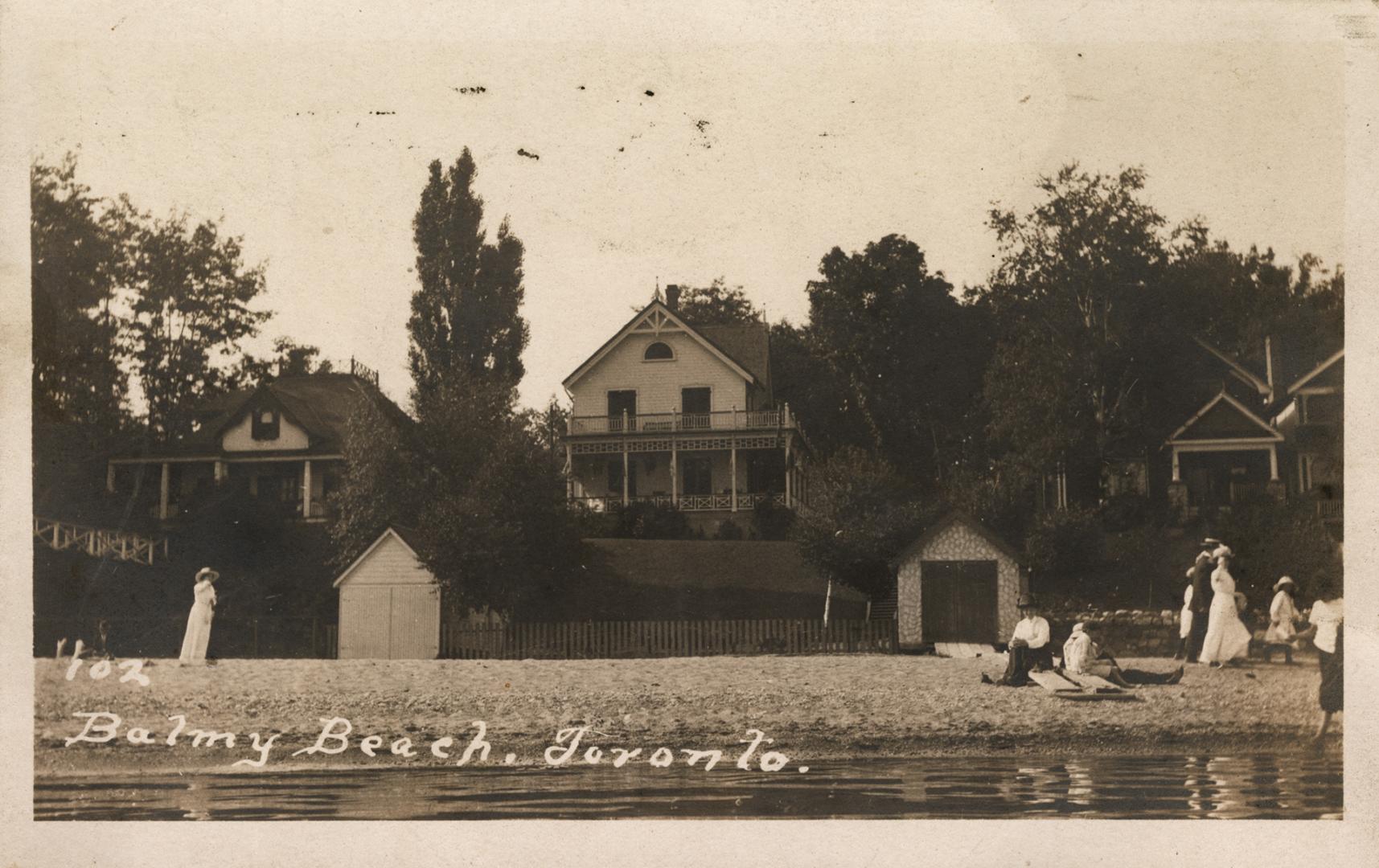 Black and white photograph of people in front of boat houses and cottages lining a sandy beach.