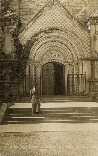 Black and white photograph of a huge, highly stylized, stone doorway. A woman is standing on th ...