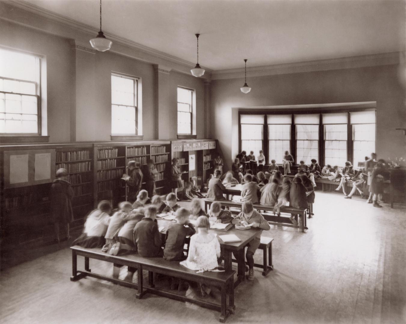 Picture of children in a large library room reading at tables and in a large window seat. 