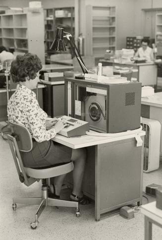 Picture of a woman working with computer equipment in a library office. 