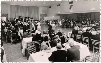 Black and white photo postcard depicting a ballroom with a band on stage and several couples da ...