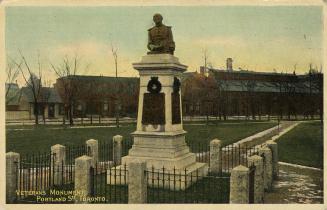 Colour photo postcard depicting a monument with a bust at the top, surrounded by a fenced-off a…