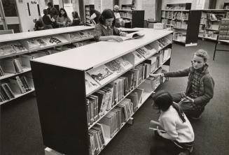 Children amongst library shelves looking at books. 