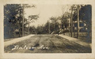 Black and white photo postcard depicting a view down an unpaved residential street with a large ...