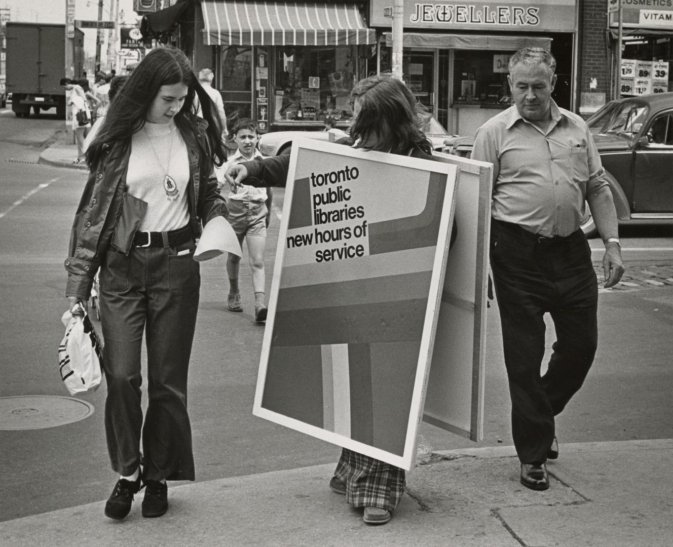 Person wearing a sandwich board advertising library hours stands on the street with two people  ...