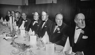 Eight older white men sit at a dinner table. Eight wear masks to resemble the face of the 8th.