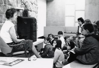 Picture of interior of library with a staff member sitting in a chair speaking to a group of ch ...