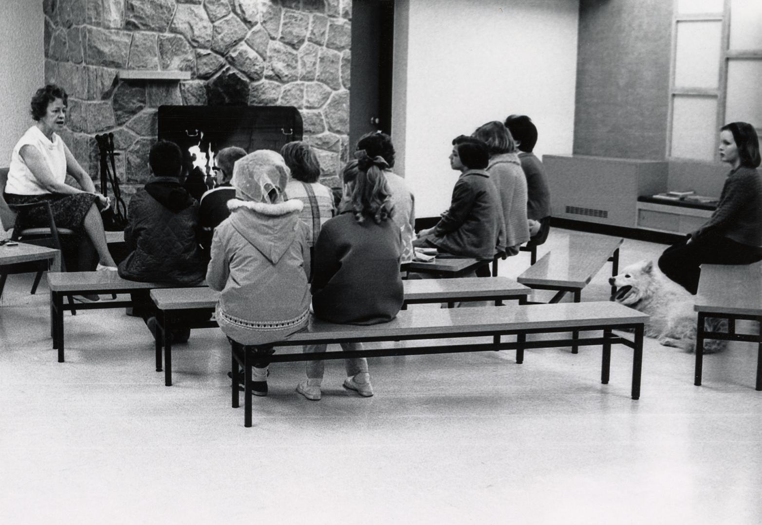 Picture of interior of library with a staff member speaking to a group of children sitting on b ...