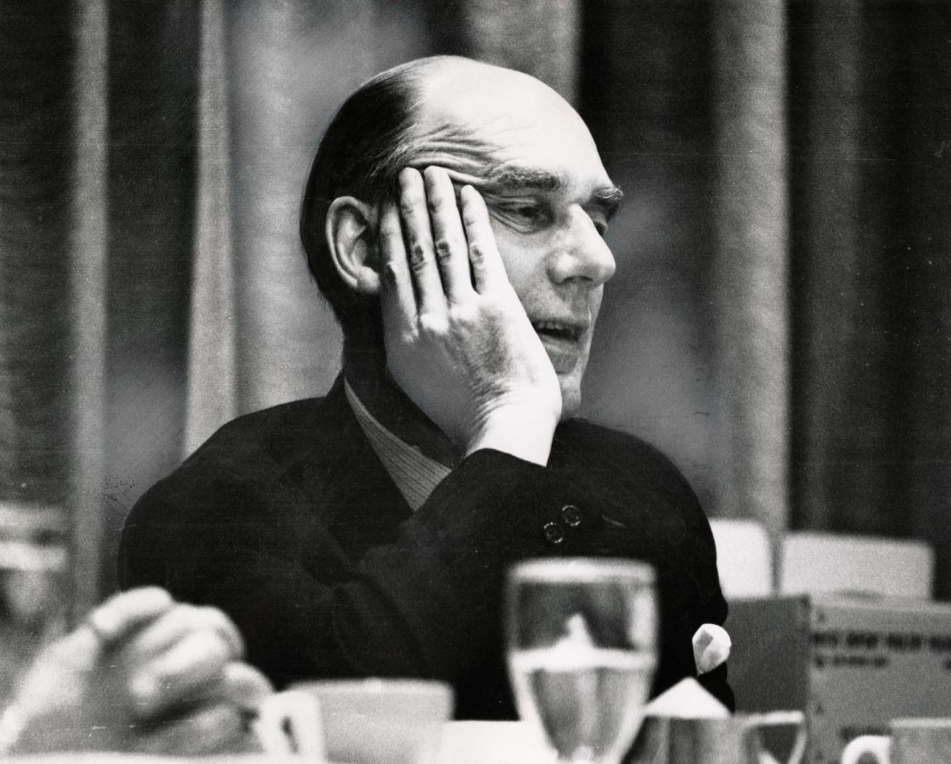 Older bald man leaning his head on his hand talking at a table with a glass of water