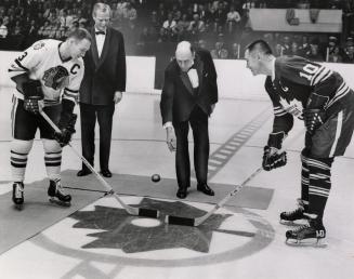 Bald white man in tuxedo drops puck between two hockey players at centre ice. A blond man in a …