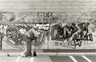 Man in safety vest with broom in front of tagged and graffiti strew wall. 