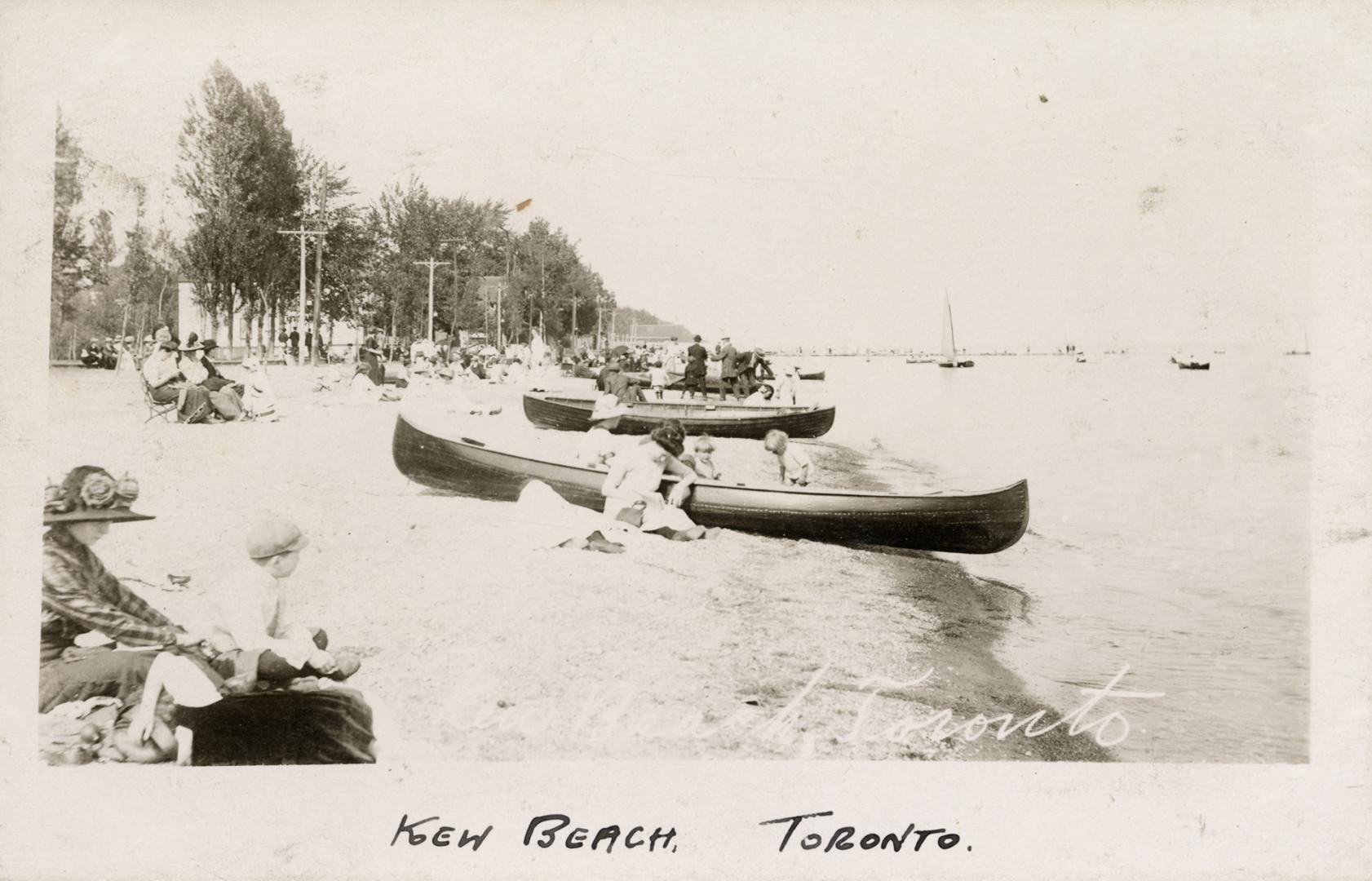 Black and white photo postcard depicting a sandy beach with canoes on the shore, people on the …