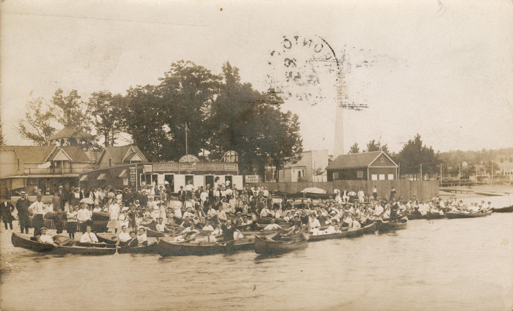 Sepia-toned photo postcard depicting many canoes filled with people along the waterfront. The b…