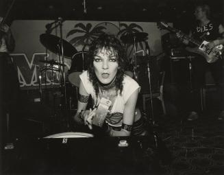 Young woman leans toward the camera in front of a drum kit in darkened room. 