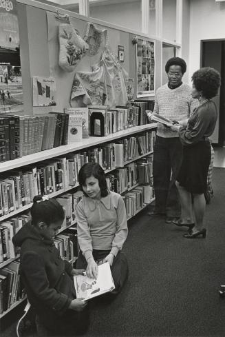 Picture of two library staff standing near shelves and two children sitting on the floor lookin ...