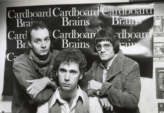 Three young stand before a poster with the text, " Cardboard Brains" repeated on it. 
