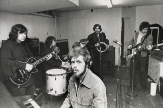 Young man at piano in foreground surrounded by four other musicians on drums, two guitars and t…