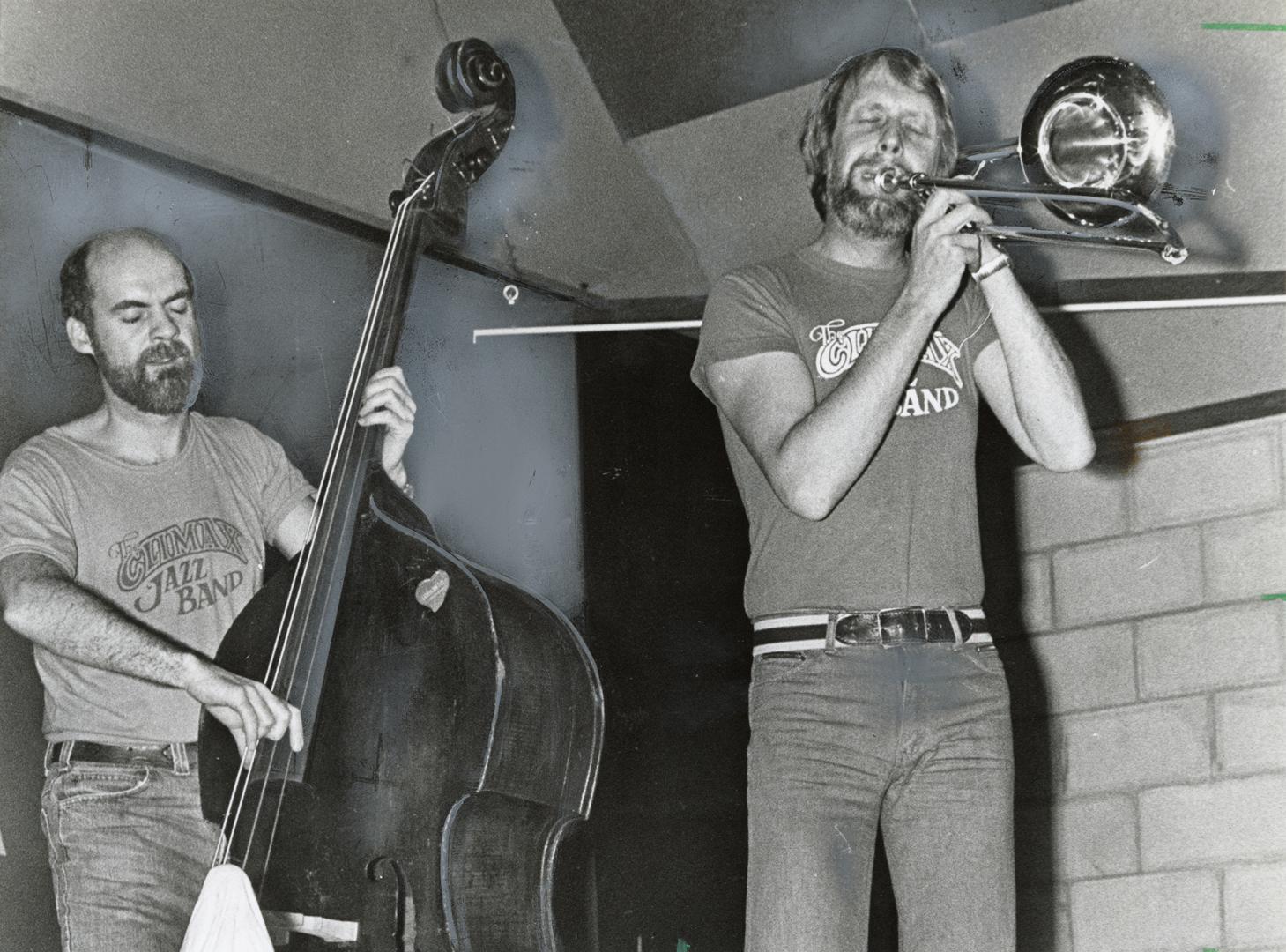 Man on bass fiddle and man playing trombone, casually dressed in t-shirts and jeans. 