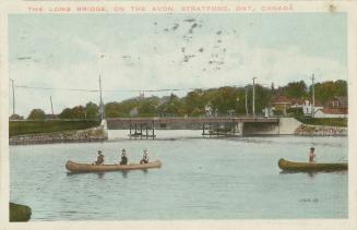 Color photograph of people in two canoes on a river with a bridge spanning it in the background…