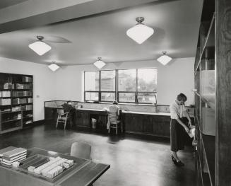 Picture of interior of a library work room with staff sitting at counter and standing at shelve…