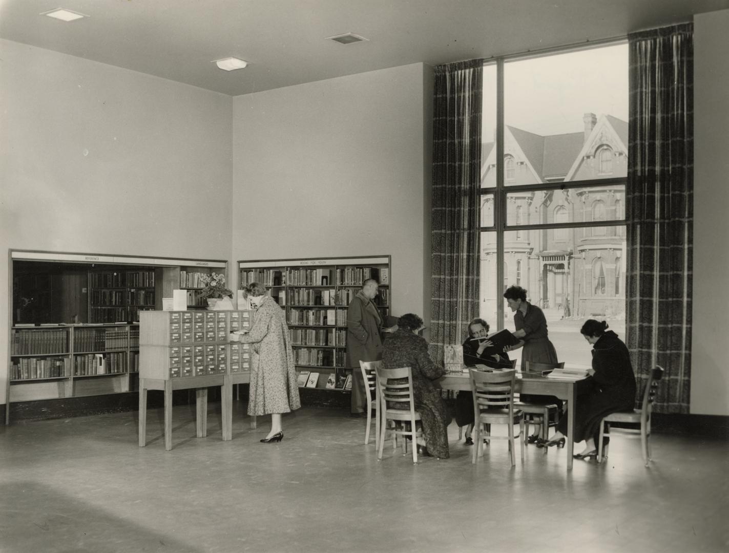 Picture of interior of a library with librarian and group of readers in front of large window. 