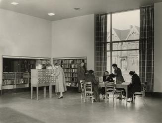 Picture of interior of a library with librarian and group of readers in front of large window. 