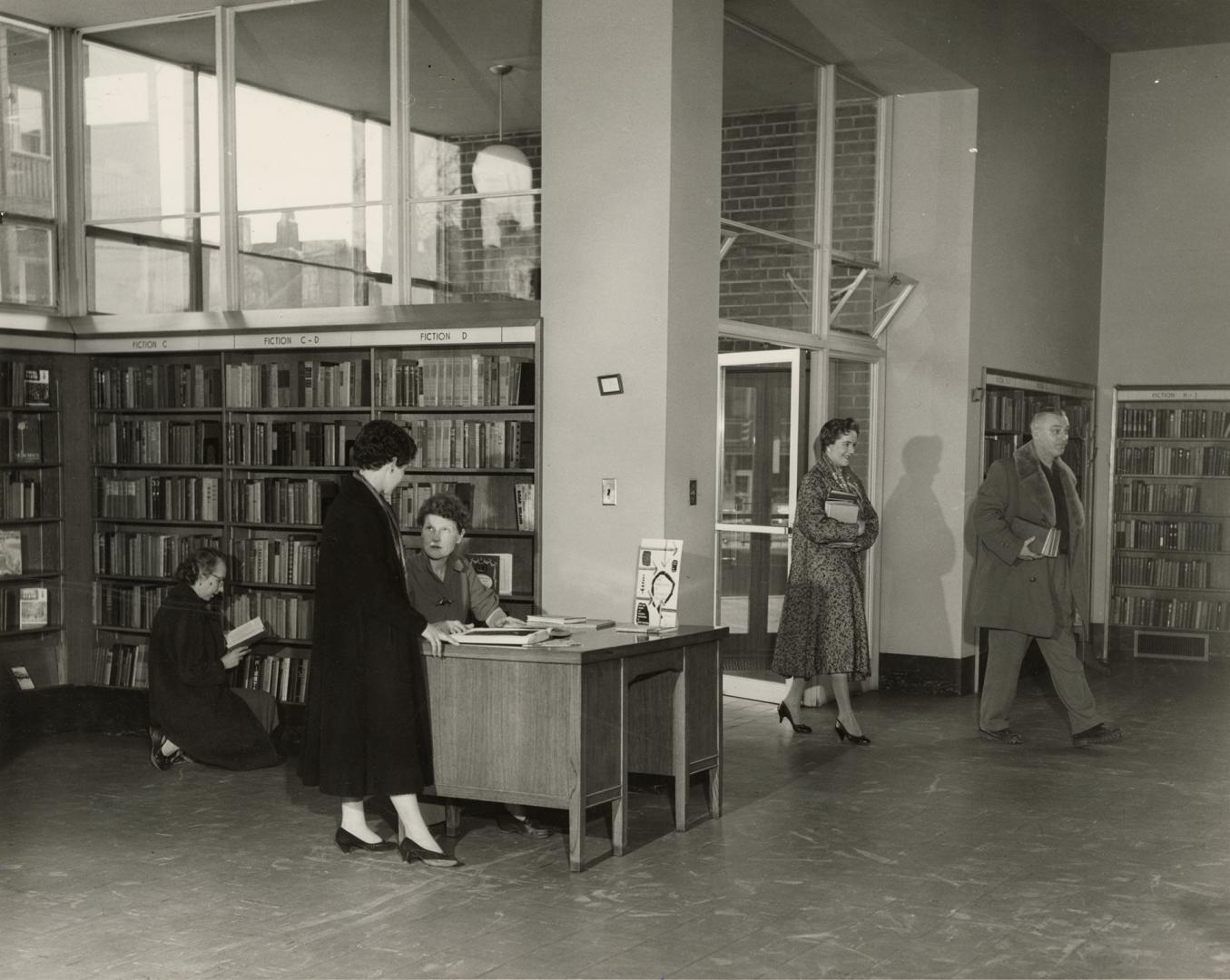 Picture of interior of a library with librarian and group of readers in front of large window. 