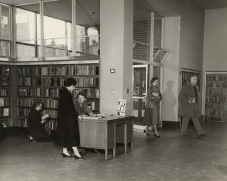 Picture of interior of a library with librarian and group of readers in front of large window. 