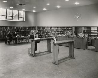 Picture of interior of a library with staff at reference desk and customers at tables. 