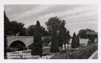 Black and white photograph of three women standing in a manicured garden with a stone bridge be…