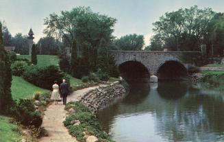 Color photograph of formal gardens with a stone walkway and a stone bridge over a river.