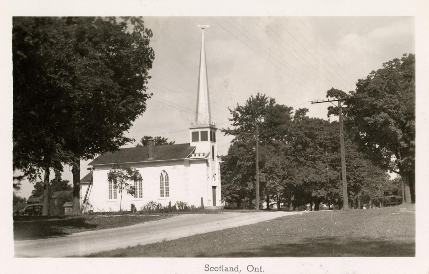 Black and white picture of a clapboard church in the gothic style by the side of a country road…